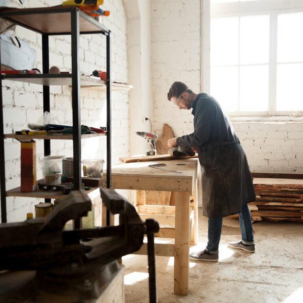 Side view of professional young joiner woodworking in planing shop garage. Skilled craftsman working on customized furniture, using sander. Carpentry services for construction, renovation and repair